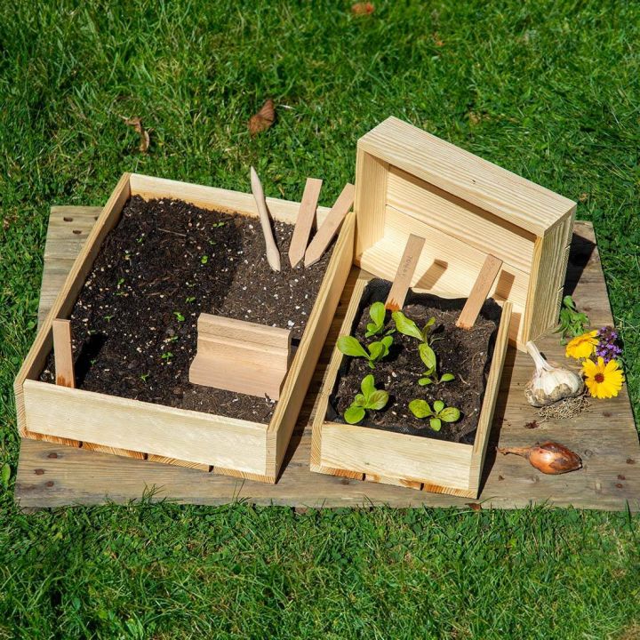 Holzk&auml;sten mit dunkler Erde, kleine gr&uuml;ne Pflanzen, Holzstecker, Gartenger&auml;te und bunte Blumen auf gr&uuml;nem Rasen.