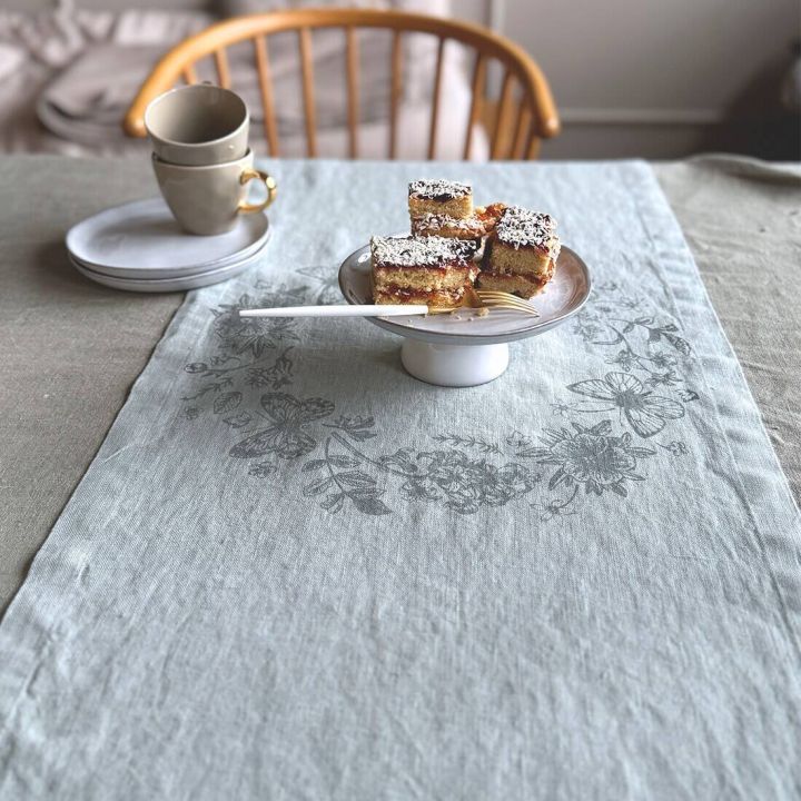 Heller Tischl&auml;ufer mit Blumenmuster, Teller mit Kuchenst&uuml;cken und Gabel, stapelweise wei&szlig;e Tassen im Hintergrund.