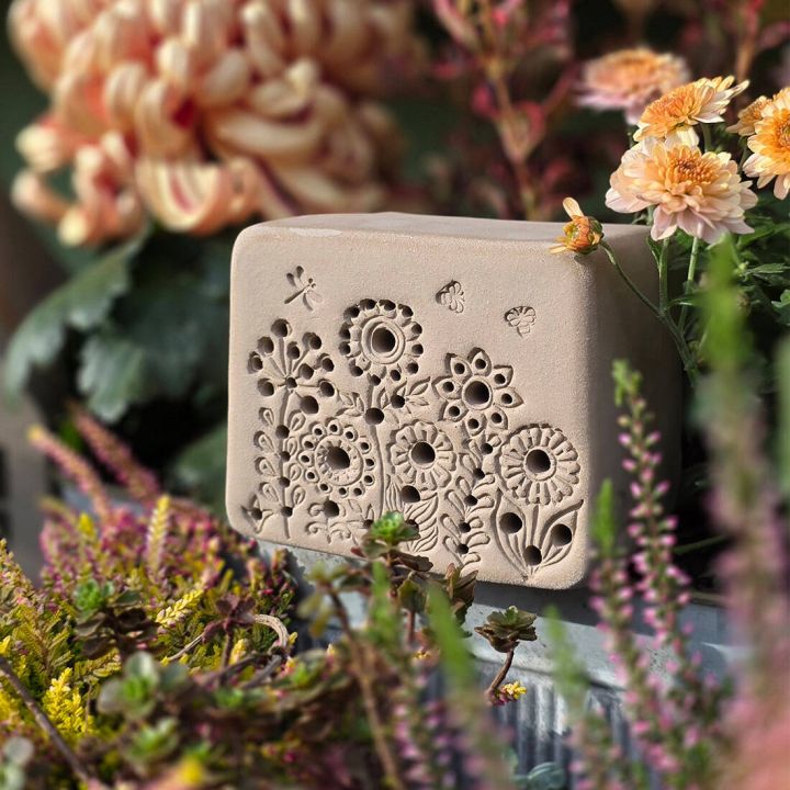 Sandfarbener Tonblock mit Blumenrelief, umgeben von bunten Blumen, weiche Formen und nat&uuml;rliche Texturen.