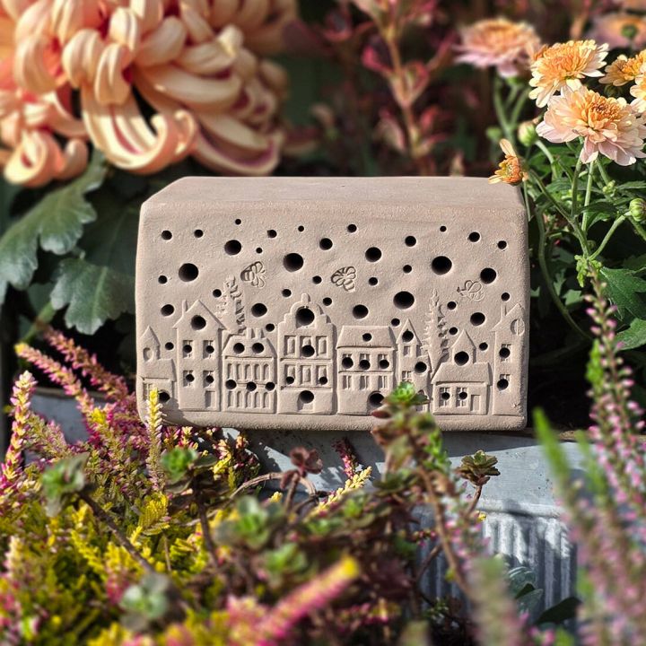 Rechteckiger, beigefarbener Tonblock mit kleinen L&ouml;chern und H&auml;userrelief, umgeben von bunten Blumen und gr&uuml;nen Bl&auml;ttern.