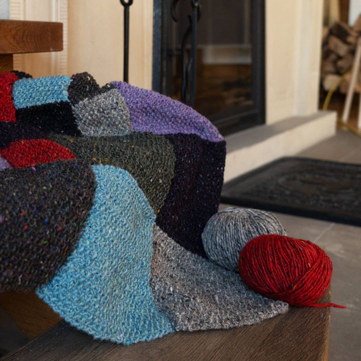 Bunte gestrickte Decke mit Patchwork-Muster, weiche Textur, daneben rote und graue Wollkn&auml;uel auf Holzboden.