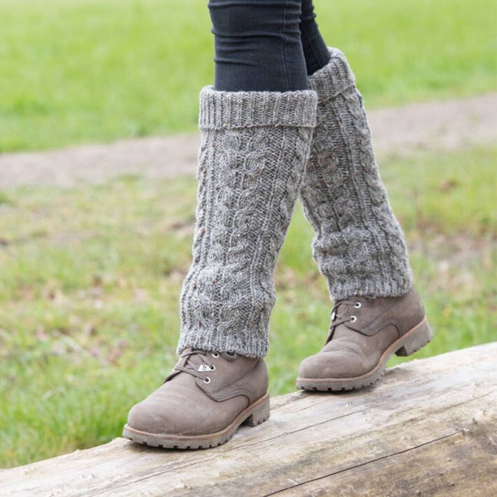 Graue, gestrickte Stulpen mit Zopfmuster, getragen &uuml;ber dunkler Hose und braunen Schn&uuml;rstiefeln, im Gr&uuml;nen auf Holzstamm stehend.