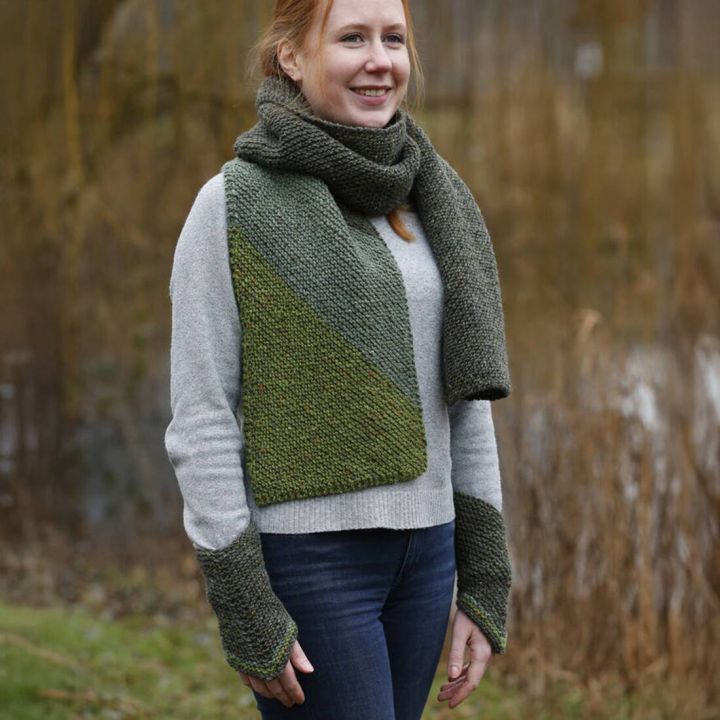 Frau mit gr&uuml;nem Schal, passenden Stulpen und grauem Pulli steht im Freien, erdige Farben, grobe Strickstruktur.