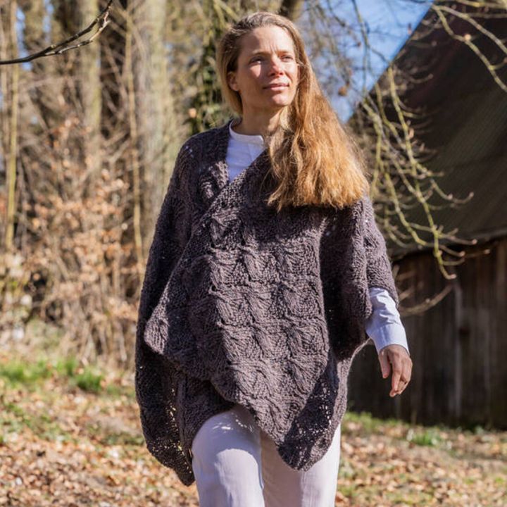 Frau mit langen blonden Haaren tr&auml;gt grob gestrickte, dunkelgraue Ponchojacke, steht im Sonnenlicht vor B&auml;umen und Holzhaus.