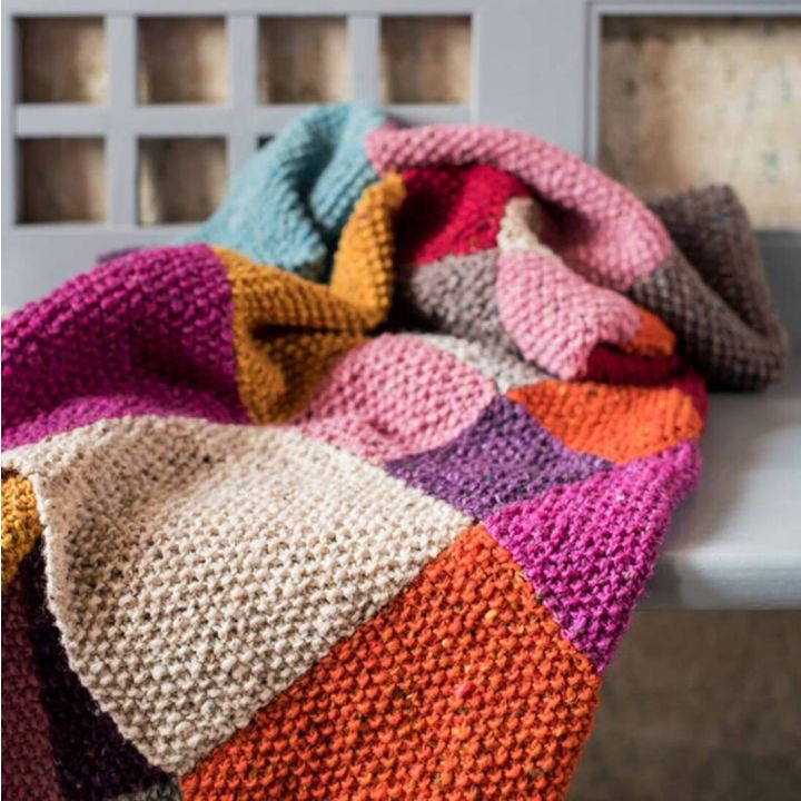 Eine gestrickte Patchworkdecke aus grobem Garn in verschiedenen Farben wie Rosa, Lila, Orange, Gelb, und Beige liegt drapiert auf einem grauen Stuhl. Die Decke besteht aus quadratischen Mustern.