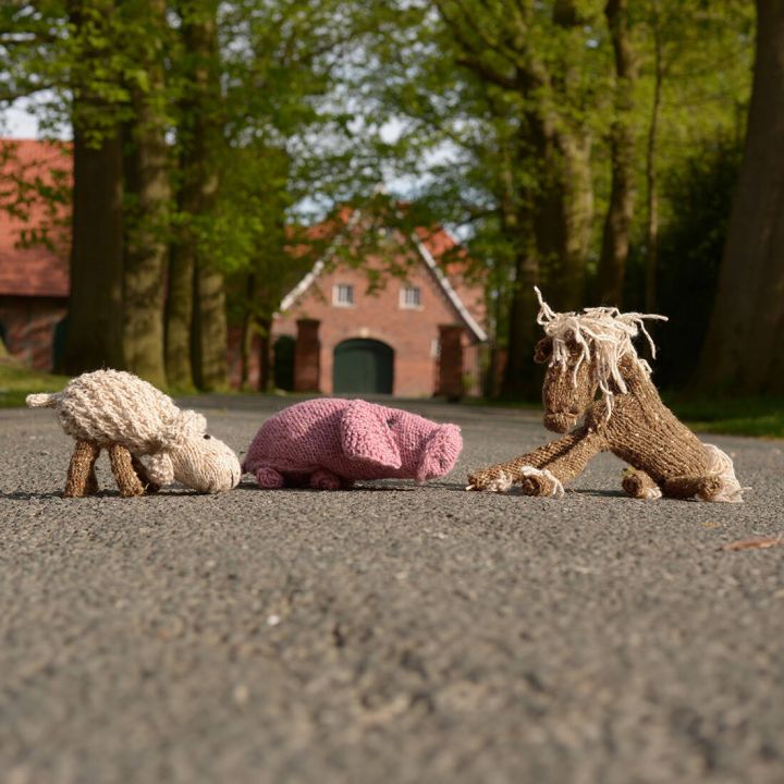 Drei gestrickte Stofftiere liegen auf einer Hofeinfahrt: ein rosa Schwein oben links, ein wei&szlig;es Schaf mit rosa Blume im Maul oben rechts und ein braunes Pferd mit heller M&auml;hne und Schweif unten im Vordergrund. Alle Tiere bestehen aus Wolle und haben