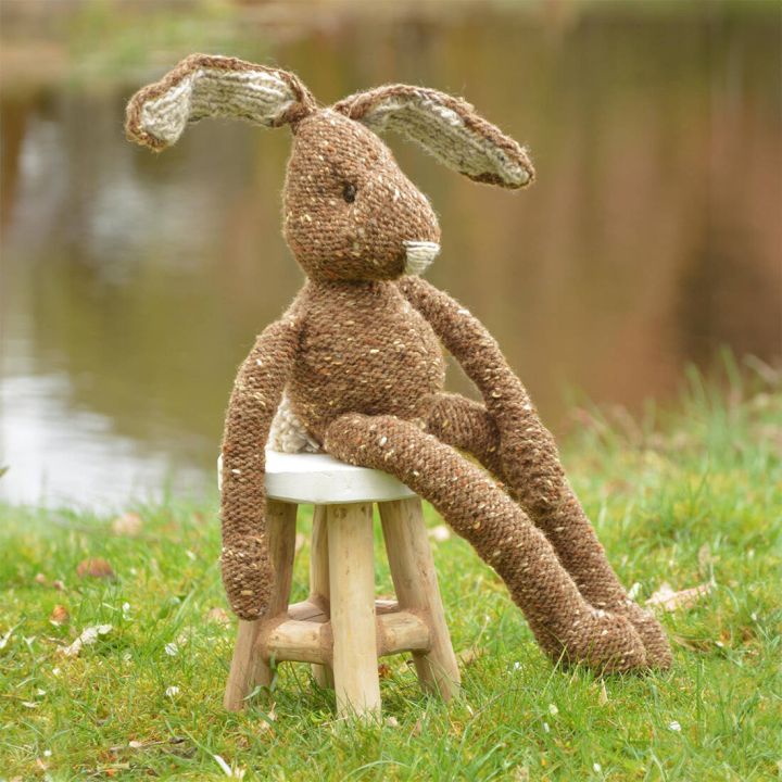 Gestrickter Hase aus brauner Wolle mit wei&szlig;en Ohren sitzt auf gr&uuml;nem Gras vor unscharfem Hintergrund.