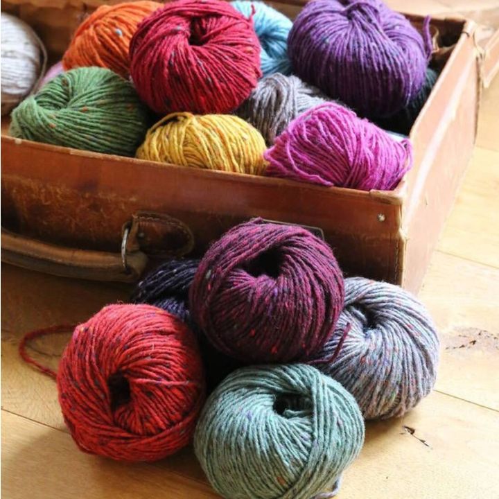 Eine Auswahl bunter Wollknäuel in verschiedenen Farben wie Rot, Gelb, Lila und Grün, liegen in einem offenen Karton auf einem Holzboden. Die Wolle ist zur Handarbeit wie Stricken oder Häkeln geeignet.