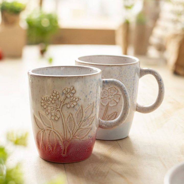 Zwei Keramiktassen mit blumenf&ouml;rmiger Pr&auml;gung und Henkel, hellgrau mit einem hellblauen Verlauf am Boden, auf einem Holztisch vor unscharfem Hintergrund mit Pflanzen.