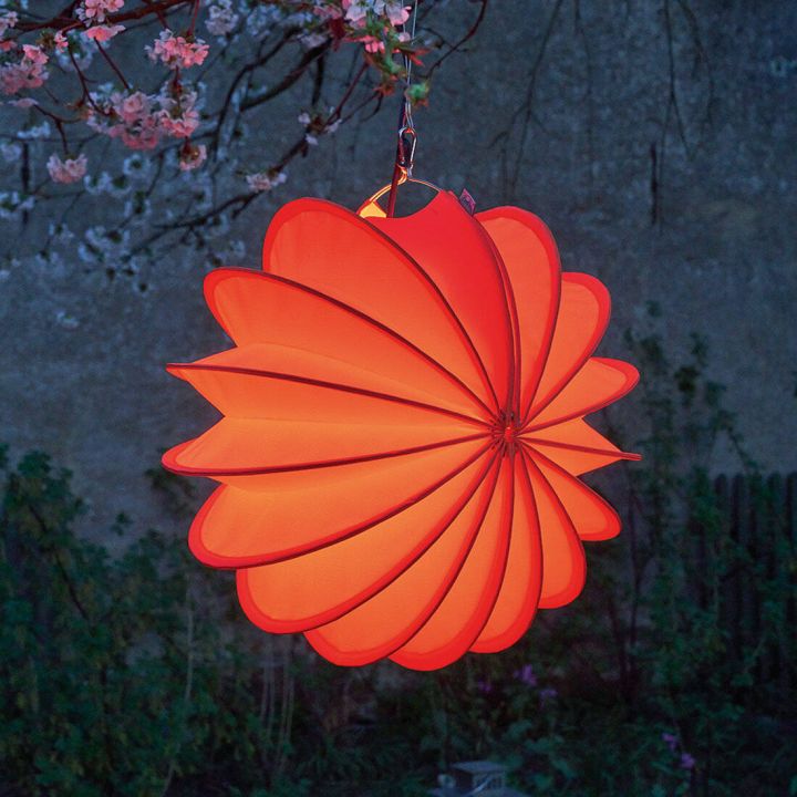 Leuchtende, orangefarbene Papierlaterne in Blütenform, aufgehängt an einem Ast mit blühenden Zweigen im Hintergrund.