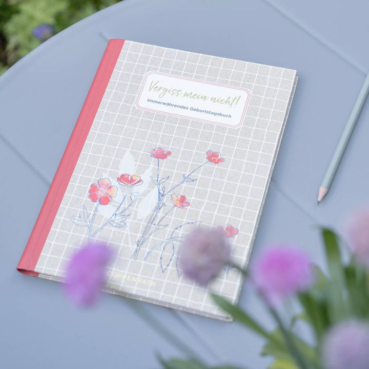 Geburtstagsbuch mit grauem, kariertem Cover, rotem Buchr&uuml;cken und Blumendesign, Titel &bdquo;Vergiss mein nicht! Immerw&auml;hrendes Geburtstagsbuch&ldquo;. Liegt auf grauem Tisch neben einem hellblauen Stift, im Vordergrund unscharfe Blumen.
