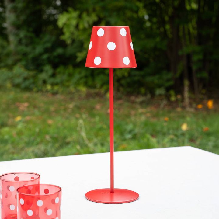 Rote Tischlampe mit wei&szlig;en Punkten, schlanker St&auml;nder, runde Basis, daneben rote Gl&auml;ser, gr&uuml;ner Hintergrund.