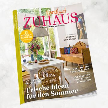 Titelbild eines Magazins „Landlust Zuhause“ zeigt einen Essbereich mit Holztisch, Korbstuhl und bunter Sitzecke. Im Hintergrund große Fenster mit Blick ins Grüne. Texte: „Frische Ideen für den Sommer“, „Wohnen mit Kunst“, „15 Sonnenschirme im Retrostil“.