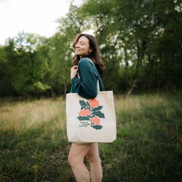 Eine Person im Freien trägt eine beigefarbene Stofftasche mit floraler Grafik und dem Text "Open eyes, open mind, open heart". Sie lächelt und steht vor Bäumen und Gras.