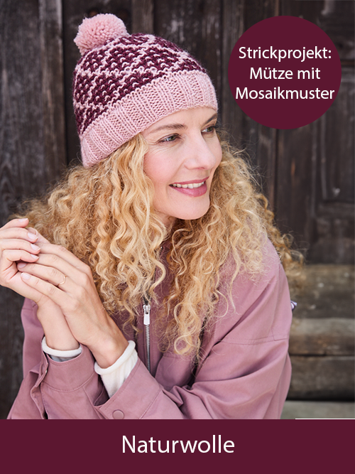 Eine lächelnde Frau trägt eine selbst gestrickte rosa-burgundfarbende Mütze. Sie ist vor einer Holzwand. Text: Strickprojekt Mütze mit Mosaikmuster. Das Bild verlinkt auf die passende Wolle: Naturwolle im Landlust Shop