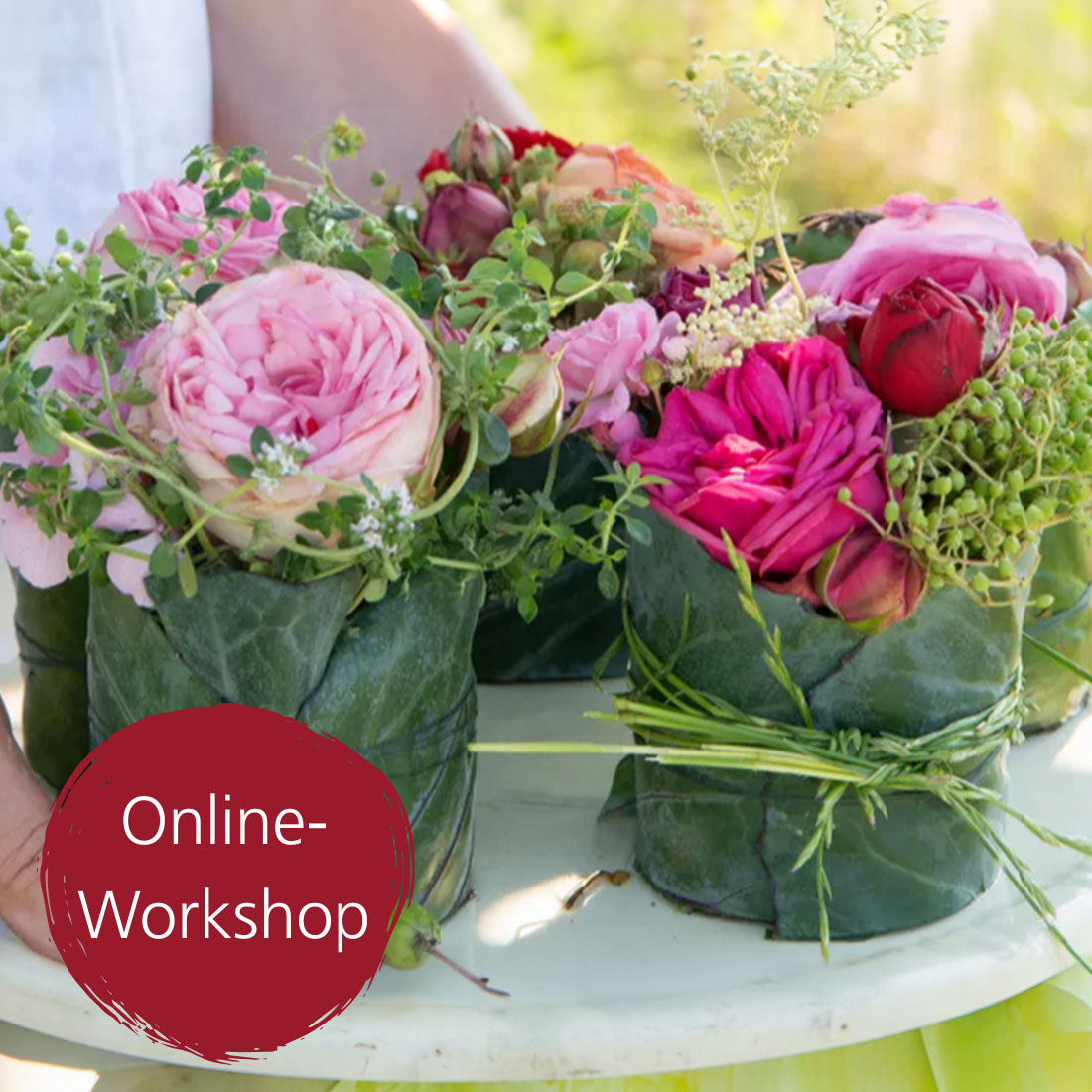 Verschiedene Blumenarrangements in gr&uuml;nen Bl&auml;ttern, bestehend aus rosa und roten Rosen, umgeben von kleinen wei&szlig;en Bl&uuml;ten und gr&uuml;nen Bl&auml;ttern, stehen auf einer Oberfl&auml;che. Bild verlinkt zum Workshop.