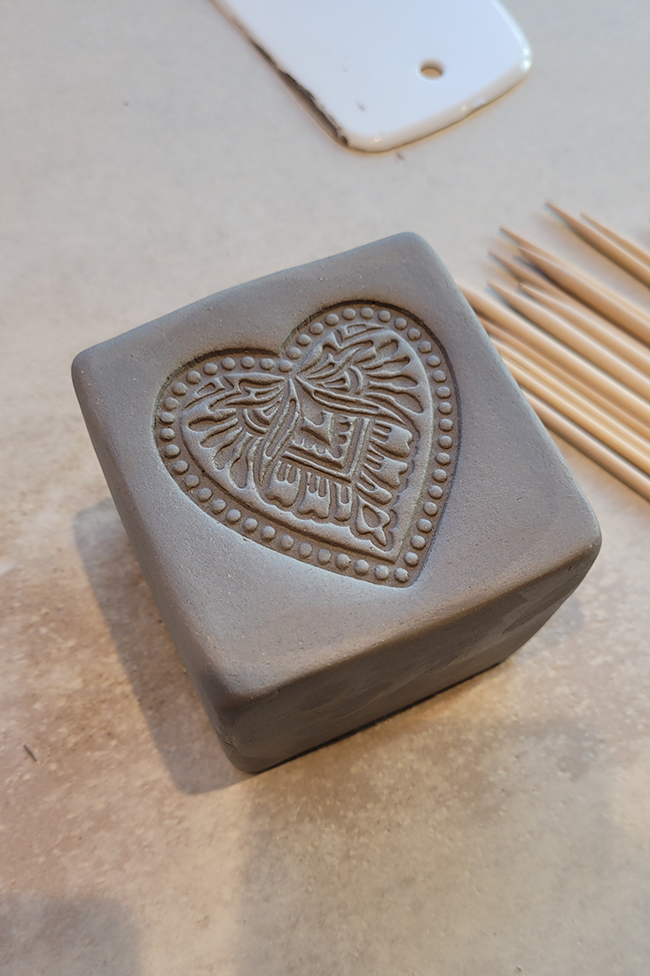 Grauer Tonw&uuml;rfel mit eingraviertem Herzmuster, glatte Oberfl&auml;che, daneben liegen d&uuml;nne Holzst&auml;bchen auf hellem Untergrund.