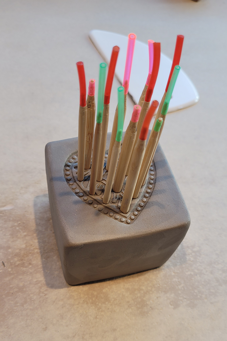 Grauer Tonblock mit Herzmuster, darin bunte St&auml;be aus Holz und Kunststoff auf hellem Untergrund.