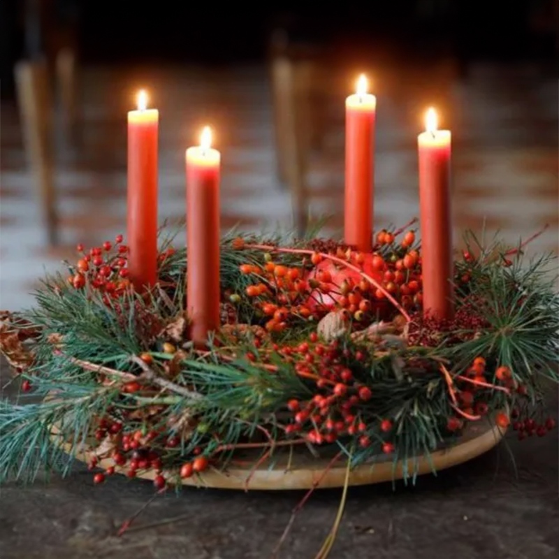 Ein Adventskranz auf Tannenzweigen und roten Beeren auf dem vier rote Kerzen brennen.