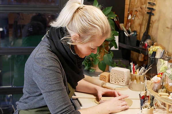 Eine Person mit blondem Haar formt Ton auf heller Arbeitsfl&auml;che, Werkzeuge und gr&uuml;ne Pflanze im Hintergrund.