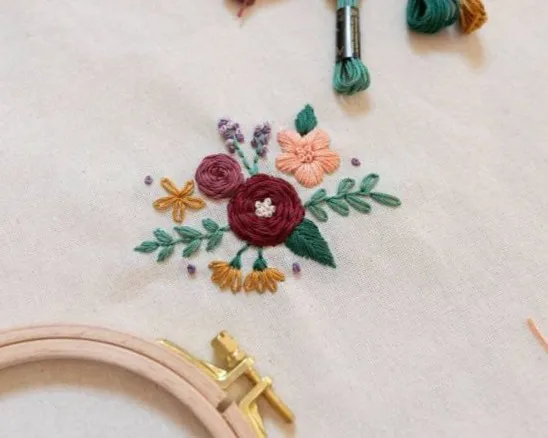 Stofftasche mit gesticktem Blumenmuster liegt auf Holztisch. Links daneben ein Stickrahmen und mehrere Bündel farbiger Stickgarne in Lila, Grün, Pink und Orange.