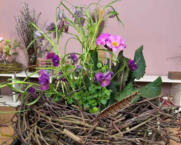 Blumenarrangement in einem Nest aus Zweigen mit violetten und rosa Blüten sowie grünen Blättern, dekoriert mit einer Feder auf rosa Hintergrund, Bild verlinkt zum Workshop.