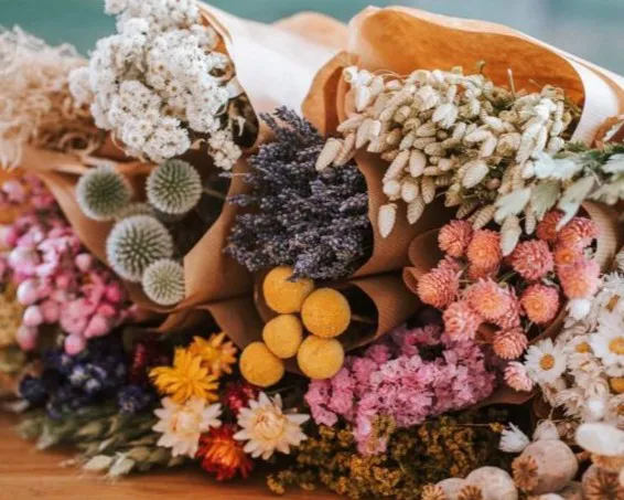 Ein Strauß aus Trockenblumen und getrockneten Gräsern in verschiedenen Farben, darunter Weiß, Lila, Rosa, Gelb und Grün, kreativ in braunem Papier arrangiert, liegt auf einem Holztisch. Bild verlinkt zum Workshop.