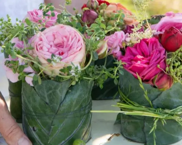 Blumensträuße in grüne Blätter gewickelt, mit Schnur gebunden. Enthalten rosa und rote Blumen, umgeben von kleinen Blättern. Hand berührt einen Strauß, Bild verlinkt zum Workshop.