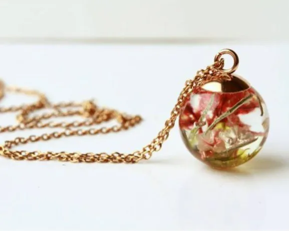 Kugelförmiger Anhänger an einer goldenen Kette, gefüllt mit getrockneten Blumen in Rot- und Grüntönen. Der Anhänger liegt auf einer weißen Oberfläche, die Kette ist locker um ihn herum drapiert. Bild verlinkt zum Produkt.