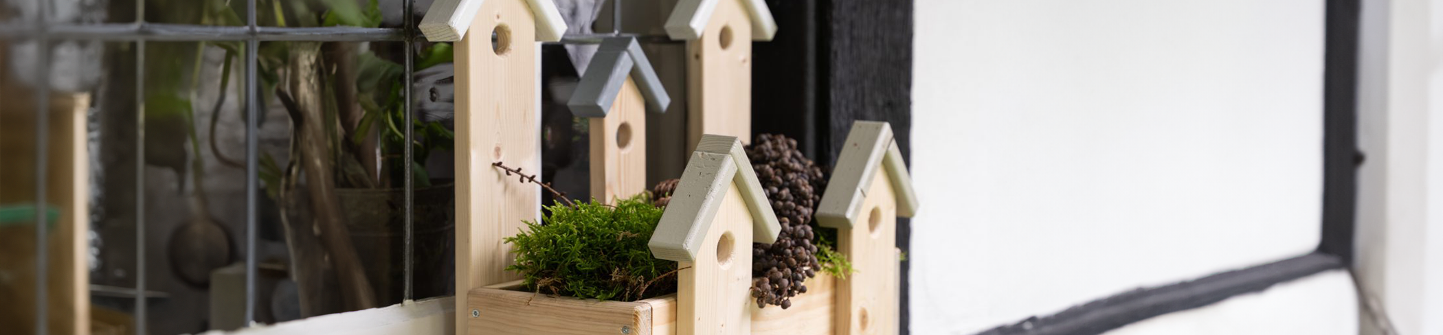 Heller Holzh&auml;userpflanzkasten mit spitzen D&auml;chern, gr&uuml;ne Pflanzen und Zapfen, schwarze und wei&szlig;e Wand im Hintergrund.