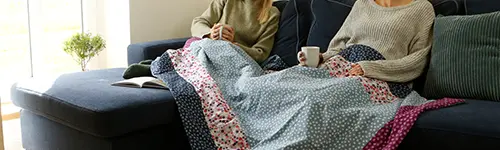 Zwei Personen sitzen in beigen Pullovern auf einem blauen Sofa, zugedeckt mit einer gemusterten Decke, halten je eine Tasse. Neben ihnen liegt ein aufgeschlagenes Buch. Im Hintergrund steht eine Topfpflanze.