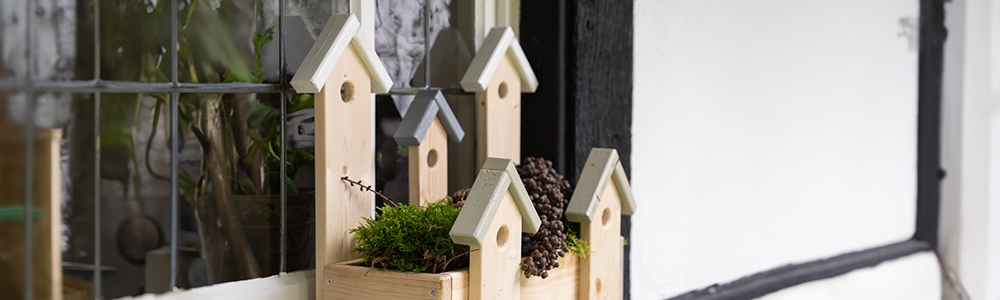 Heller Holzh&auml;userpflanzkasten mit spitzen D&auml;chern, gr&uuml;ne Pflanzen und Zapfen, schwarze und wei&szlig;e Wand im Hintergrund.