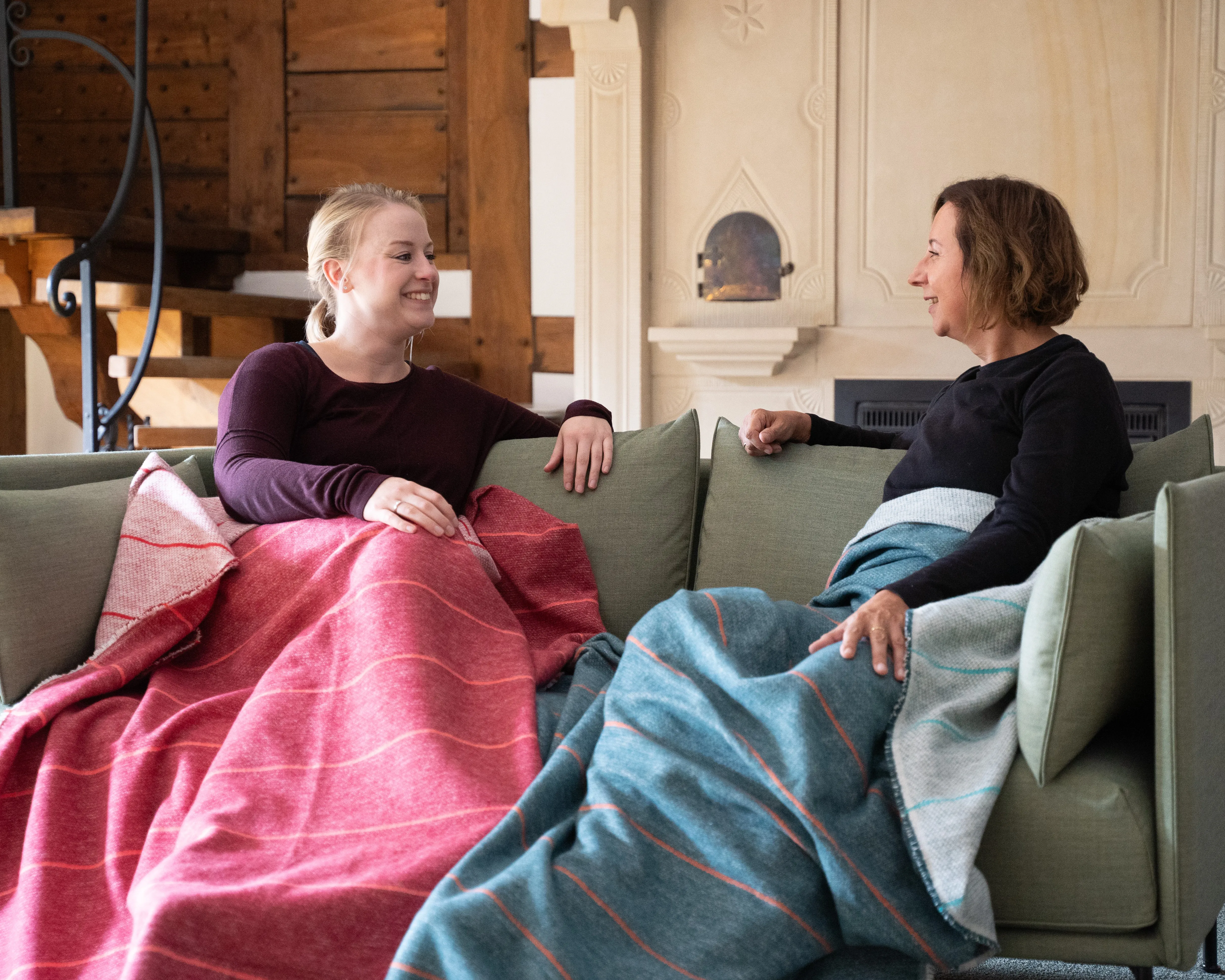 Zwei Frauen sitzen auf einem Sofa und unterhalten sich, eingehüllt in bunte Decken. Links trägt eine Frau eine rote Decke, rechts eine Frau eine blaue Decke. Im Hintergrund sind eine Kaminverkleidung und Holzelemente sichtbar. Bild verlinkt zu Landingpage