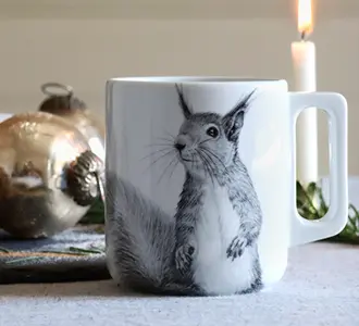 Auf einer weißen Tasse ist ein schwarz-weiß gezeichnetes Eichhörnchen. Im Hintergrund befindet sich eine Kerze und Weihnachtskugeln.