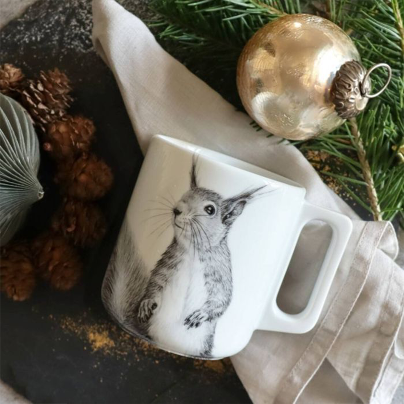Auf einer weißen Tasse ist ein schwarz-weiß gezeichnetes Eichhörnchen. Im Hintergrund befindet sich eine Kerze und Weihnachtskugeln.