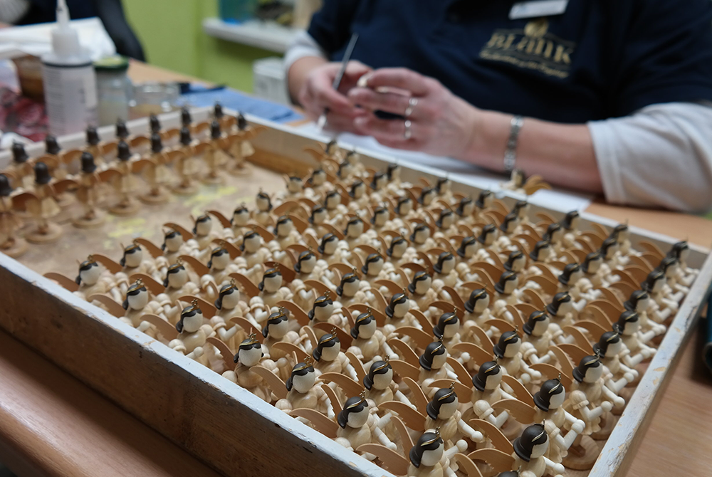 Eine Person bemalt kleine Engelfiguren aud Holz. Die Figuren stehen alle ordnentlich aufgereiht auf einem Tablett