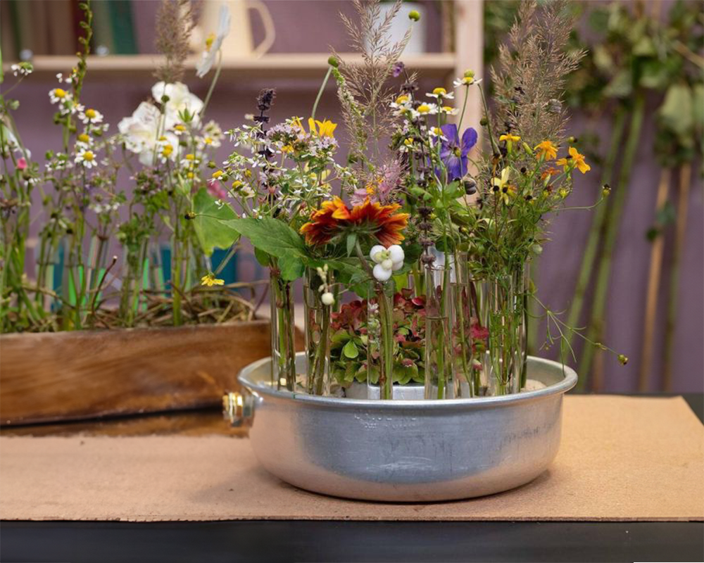 Ein Blumengesteck in einer metallenen Schale steht auf einem Holztisch. Im Hintergrund sind weitere Pflanzen und Gefäße zu erkennen.