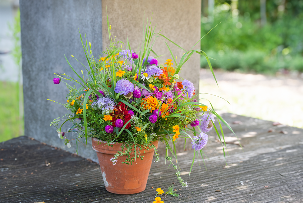 Ein bunter Blumenstrauß steht in einem braunen Topf. Dieser ist im Außenbereich auf einem Holzboden.