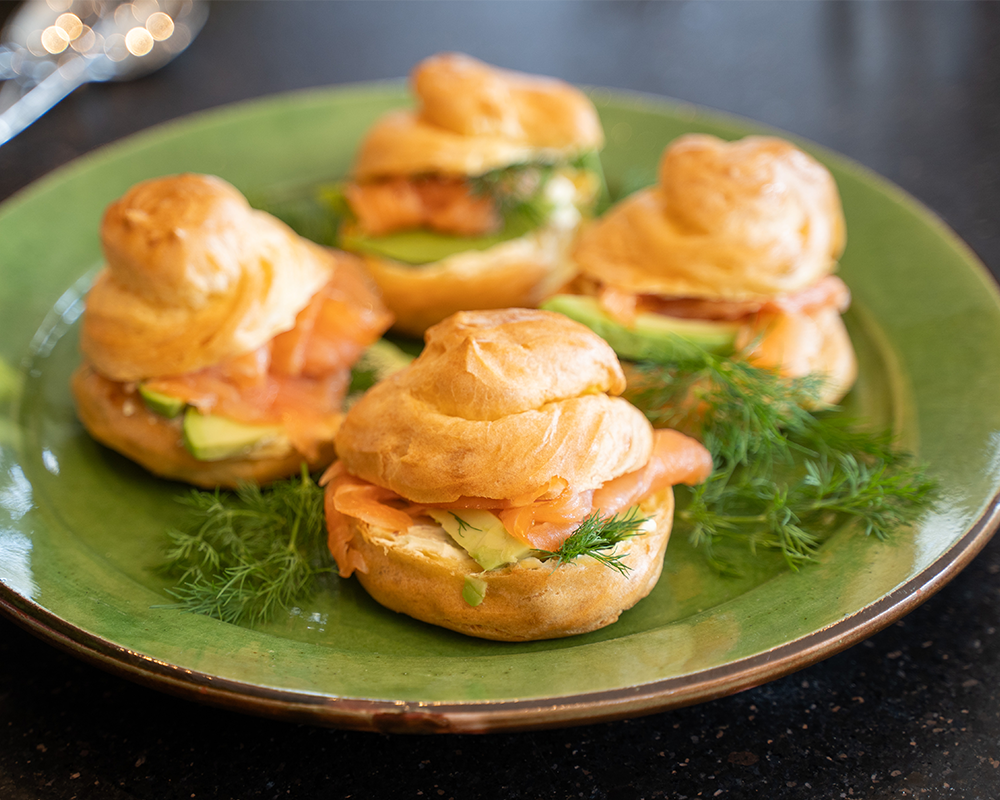 Auf einem grünen Teller sind vier kleine Sandwiches aus Brandteig mit Lachs gefüllt