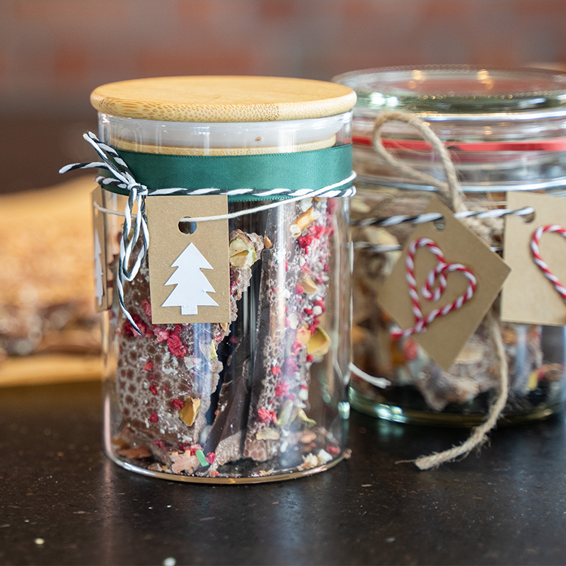 Zwei Einmachgläser mit bunt verzierter Bruchschokolade gefüllt. Sie sind dekoriert mit weihnachtlichen Anhängern