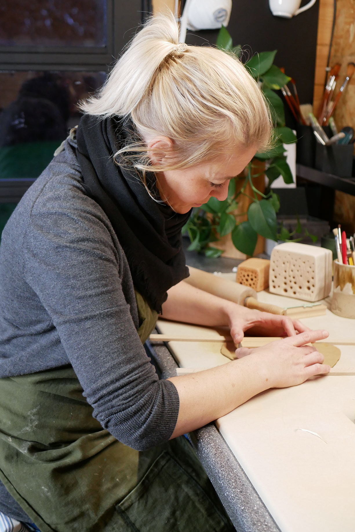 Frau mit blondem Haar formt Ton an einem Tisch, gr&uuml;ne Pflanze im Hintergrund, warme Farben, kreative Atelier-Atmosph&auml;re.