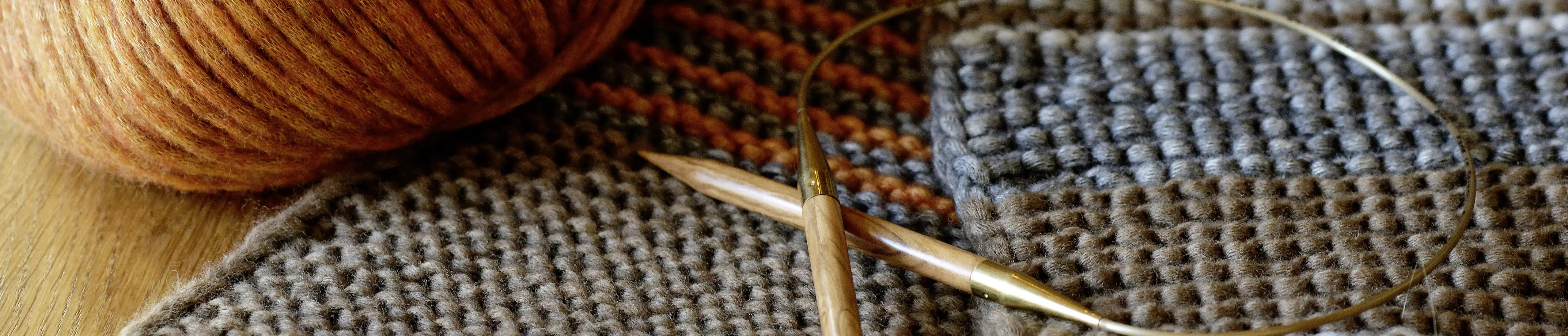 Eine Stricknadel aus Holz liegt neben einem orange-braunen Wollkn&auml;ul auf einem gestrickten Schal.