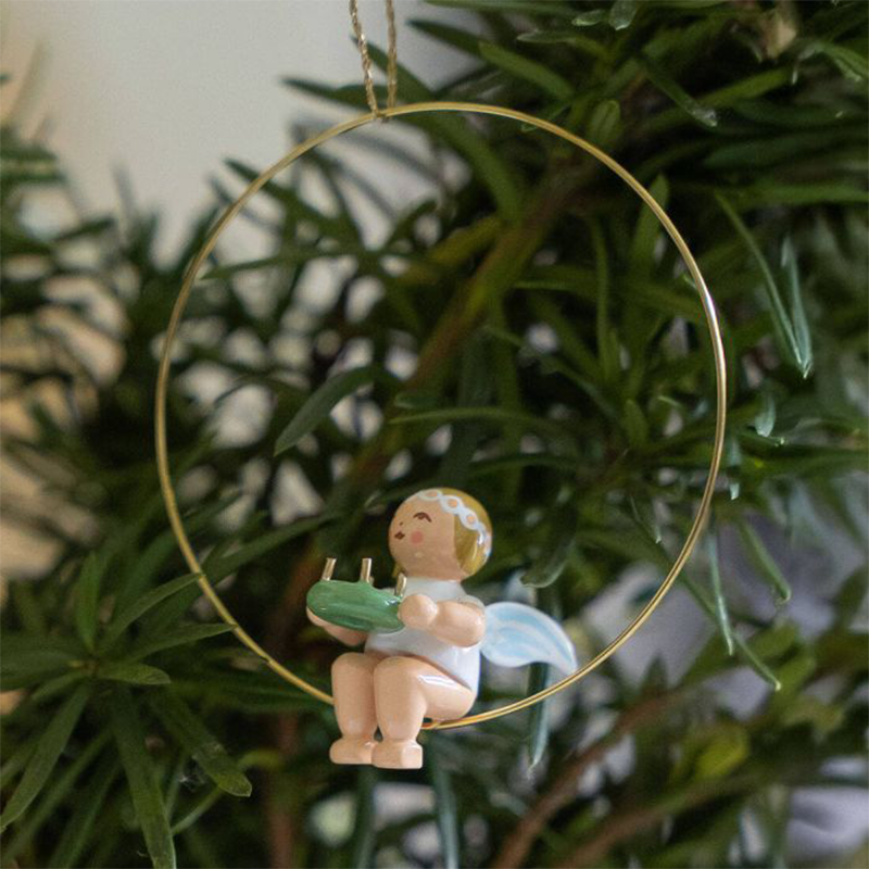Eine Figur in Form eines Engels sitzt einem goldenen Ring. Dieser hängt an einem Tannenzweig