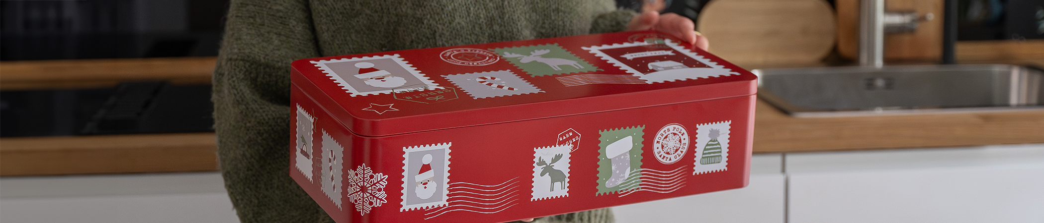 Eine extragroße Gebäckdose in Rot mit weihnachtlichen Motiven wird von einer Person mit grünem Oberteil gehalten