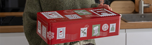 Eine extragroße Gebäckdose in Rot mit weihnachtlichen Motiven wird von einer Person mit grünem Oberteil gehalten