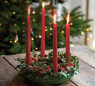 Ein Kranz aus grünem Keramik auf dem vier rote Stabkerzen brennen. Er ist mit Tannenzweigen und Beeren geschmückt. Im Hintergrund ist ein Weihnachtsbaum zu erkennen.