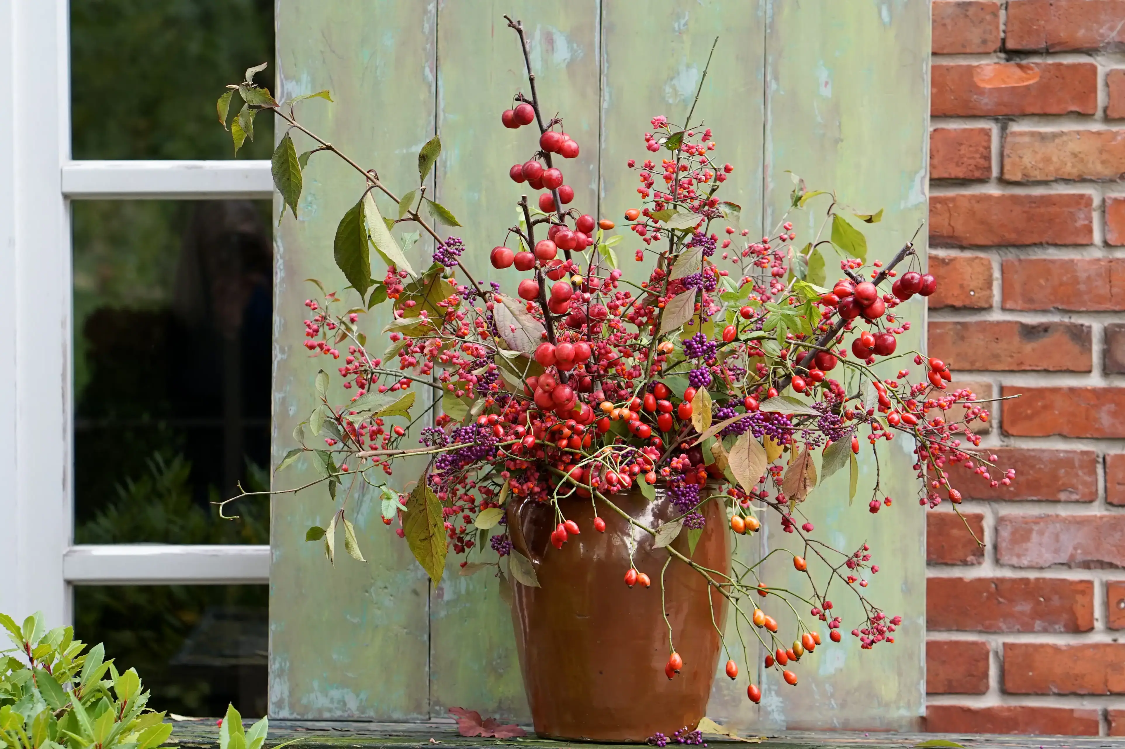 Pflanzen wie Hagebutten, &Auml;ste mit Bl&auml;ttern etc. stehen gesteckt in einem rost-braunen Blumentopf vor einer Hauswand.