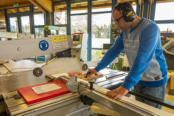 Ein Mann trägt Gehörschutz und arbeitet in einer Holzwerkstatt. Er bedient eine große Säge, um ein Holzstück zu schneiden. Der Raum hat große Fenster, durch die Tageslicht einfällt. Bild verlinkt zum Herstellerportrait. 