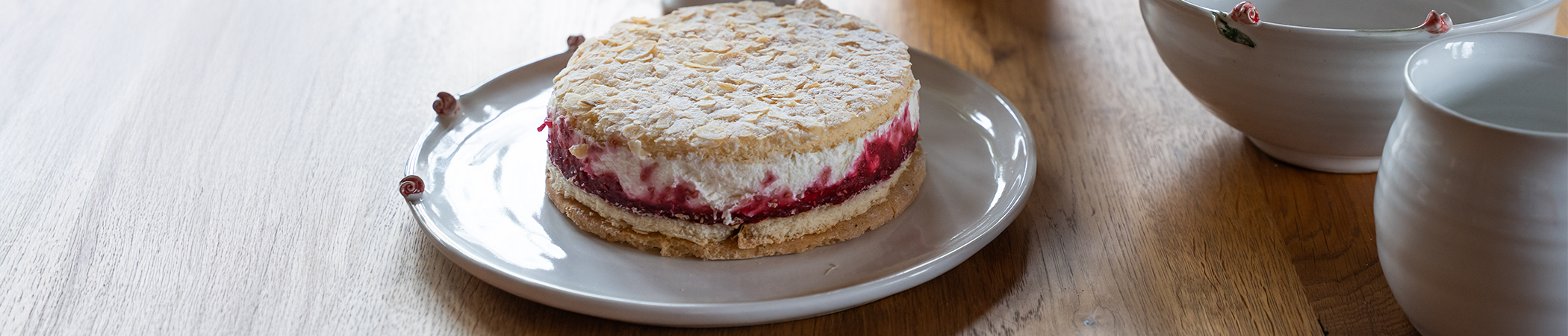 Eine Torte mit Beeren und Sahne auf einer weißen Tortenplatte steht auf einem Holztisch. Drum herum ist weitere weiße Keramik