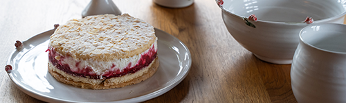 Eine Torte mit Beeren und Sahne auf einer weißen Tortenplatte steht auf einem Holztisch. Drum herum ist weitere weiße Keramik
